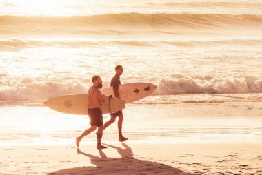 Two surfers carry their boards along the sandy shore during a vibrant sunset in Arraial do Cabo.